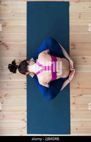 Vista dall'alto della giovane donna caucasica seduta in dormiente tartaruga posa suppta kurmasana sul pavimento in studio. Foto Stock