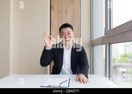 ritratto di giovane asiatico felice lavoratore maschile ufficio guardando la macchina fotografica e sorridendo in interni. Primo piano di un manager amichevole in un abito formale e bianco shir Foto Stock