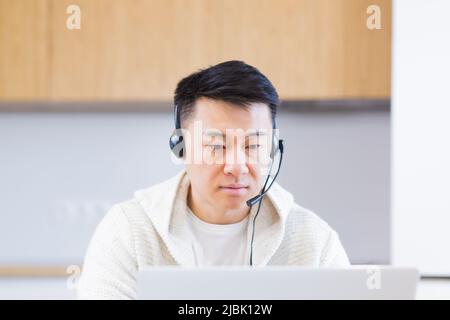 giovane uomo asiatico freelance seduto a casa alla scrivania con cuffia e parlare online con collegamento video via computer portatile in camera o in cucina. Il maschio interno Foto Stock