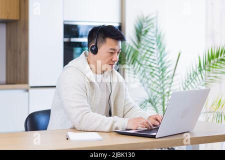 giovane uomo asiatico freelance seduto a casa alla scrivania con cuffia e parlare online con collegamento video via computer portatile in camera o in cucina. Il maschio interno Foto Stock