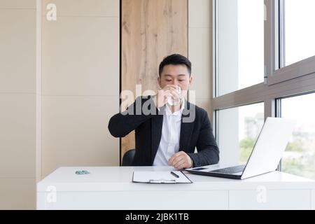 giovane lavoratore asiatico ufficio che soffre di mal di testa al lavoro in ufficio bevande pillola medicina con acqua. Uomo malato in un vestito al computer interno wi Foto Stock