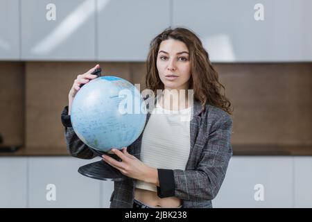 ritratto di una giovane insegnante di sesso femminile che insegna una lezione di geografia online da remoto, tenendo un globo, puntando verso un globo di paese, guardando la fotocamera Foto Stock