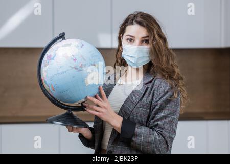 ritratto di una giovane insegnante femminile in una maschera medica insegnamento di una lezione di geografia online da remoto, tenendo un globo, puntando verso un globo globale, lookin Foto Stock