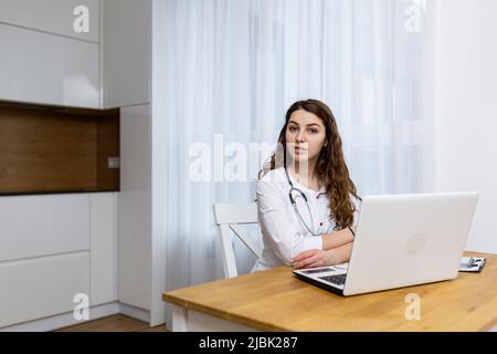 Giovane dottore donna tratta online da casa, si consulta attraverso una videochiamata da un computer portatile, seduto in cucina in un cappotto bianco, guardando il camer Foto Stock