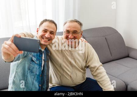 Sorridente figlio adulto sedersi sul divano rilassarsi con padre senior parlare condividere pensieri guardando negli occhi, felice uomo di riposo millenario sul divano parlare con gli anziani Foto Stock