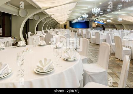 Sala banchetti decorata in bianco con tavoli rotondi serviti e palco per lo spettacolo Foto Stock