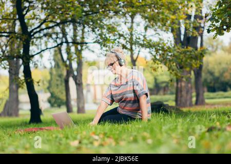 Bella donna bionda, anziana, seduta sull'erba nel parco in cuffie, utilizza un computer portatile, opere, studi, ascolta musica, parla del video pl Foto Stock