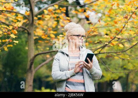 Bella donna anziana nel parco a piedi utilizza il telefono, parlando al telefono, sorridendo Foto Stock