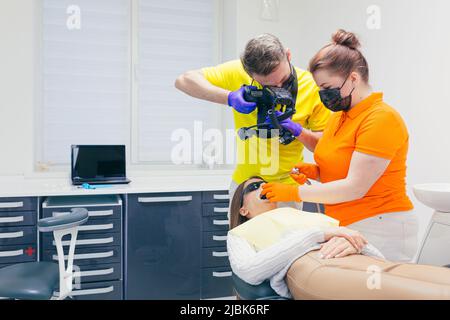 Un dentista tratta i denti di una giovane donna e un assistente le fotografa i denti. Sdraiato in una sedia dentale in ufficio Foto Stock