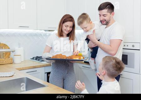 Colazione per tutta la famiglia, mamma, papà, due bambini, ragazzi che mangiano croissant appena cotti da mamma, caffè e succo di frutta fresco, sorridendo, abbracciando a casa nella cannella Foto Stock