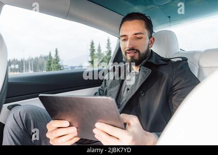 Giovane caucasico uomo di successo che parla seriamente sullo smartphone mentre si siede sul sedile posteriore di un'auto di guida Foto Stock