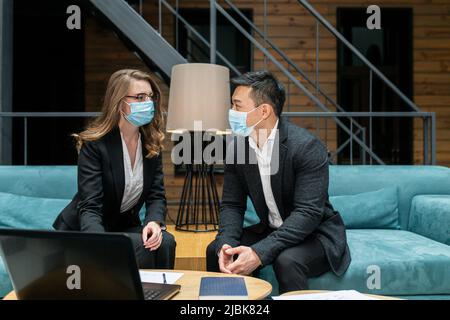 Due dipendenti di un'azienda internazionale discutono di un rapporto sul lavoro svolto, un uomo asiatico e una donna che indossa maschere mediche protettive, seduto nel Foto Stock