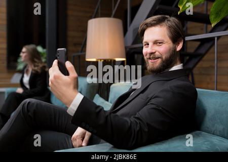 Uomo d'affari maschile seduto in un elegante ristorante, in un abito business rilassante, e utilizza il telefono per comunicare Foto Stock