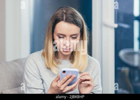 giovane donna felice ha visto le buone notizie su smartphone, in piedi accanto alla finestra in ufficio o a casa al chiuso. Femmina con delizia tiene un telefono cellulare in hi Foto Stock