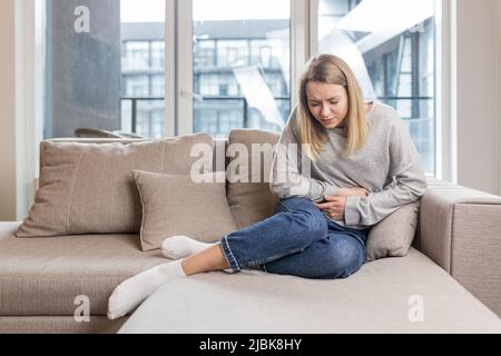 Una giovane donna è seduta sul divano a casa, tenendo in mano uno stomaco che fa male, urla nel dolore, sente dolore, è malata, si sente male Foto Stock