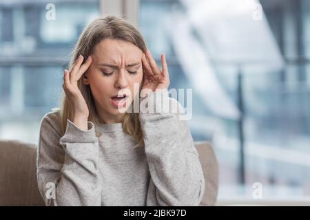 La giovane donna ha un mal di testa, tiene la testa con le mani e mostra dolore a casa, in ufficio al lavoro Foto Stock