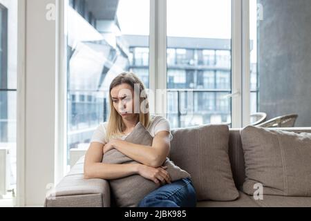 Giovane donna turbata seduta sul divano a casa, putting cuscino, sensazione di cattivo umore, depresso, cattivo umore Foto Stock