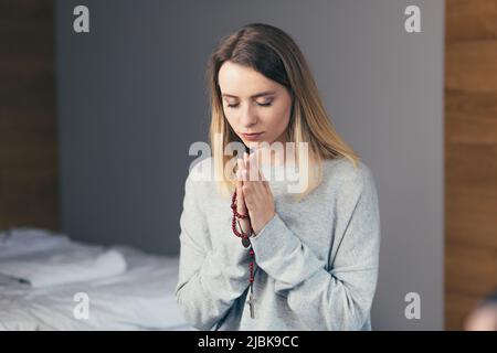 Ritratto di una giovane donna che ha piegato le braccia e pregato, tenendo un rosario nelle mani, con gli occhi chiusi, calmo Foto Stock