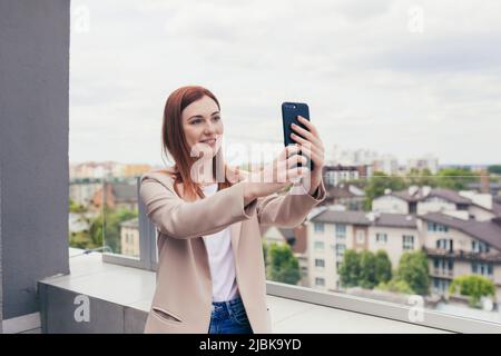 Giovane bella donna, lavoratore d'ufficio, sorridente e utilizza il telefono per la comunicazione video, donna d'affari sul balcone del centro d'ufficio Foto Stock