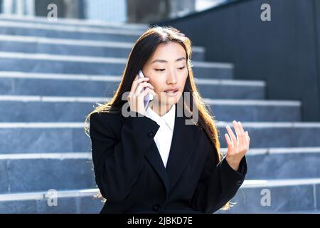 Donna asiatica seduta triste sulle scale di un edificio moderno segnala cattive notizie in una conversazione telefonica, sconvolto e depresso Foto Stock