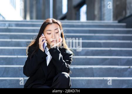 Donna asiatica seduta triste sulle scale di un edificio moderno segnala cattive notizie in una conversazione telefonica, sconvolto e depresso Foto Stock