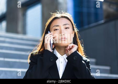 Donna asiatica seduta triste sulle scale di un edificio moderno segnala cattive notizie in una conversazione telefonica, sconvolto e depresso Foto Stock