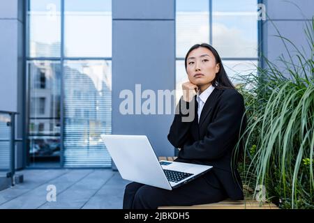 Donna d'affari triste e pensiva che lavora su un portatile seduto su una panchina vicino all'ufficio, asiatico stanco e depresso in cerca di lavoro Foto Stock