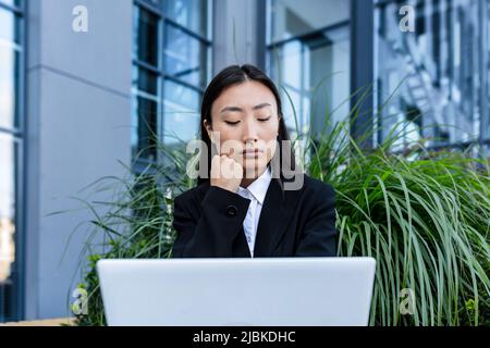 Donna d'affari triste e pensiva che lavora su un portatile seduto su una panchina vicino all'ufficio, asiatico stanco e depresso in cerca di lavoro Foto Stock