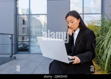 Donna d'affari triste e pensiva che lavora su un portatile seduto su una panchina vicino all'ufficio, asiatico stanco e depresso in cerca di lavoro Foto Stock