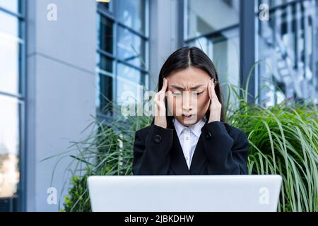 Donna d'affari triste e pensiva che lavora su un portatile seduto su una panchina vicino all'ufficio, asiatico stanco e depresso in cerca di lavoro Foto Stock