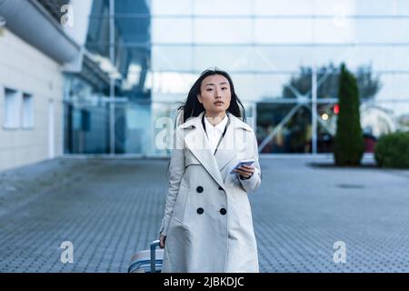 Bella donna turistica vicino all'aeroporto, una donna cinese che cammina con una grande valigia, una donna asiatica che tiene un telefono, utilizzando la domanda di libro Foto Stock