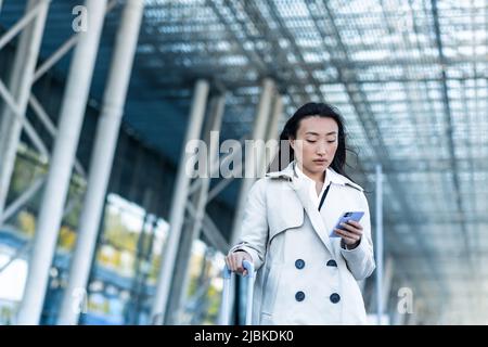 Bella donna turistica vicino all'aeroporto, una donna cinese che cammina con una grande valigia, una donna asiatica che tiene un telefono, utilizzando la domanda di libro Foto Stock