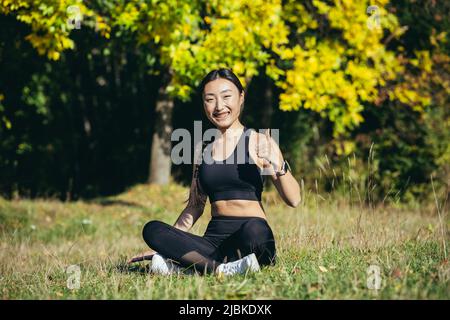 Bella donna asiatica medita nei boschi in una giornata di sole, l'atleta si riposa seduto in posizione lotus e guarda felicemente e con calma al camer Foto Stock