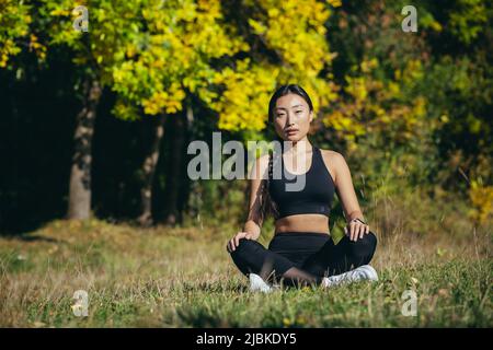 Giovane bella sport ragazza asiatica donna meditating in parco, seduta lotus posa praticare yoga mat, zen. Rilassa all'aperto in natura nel mattino con Foto Stock