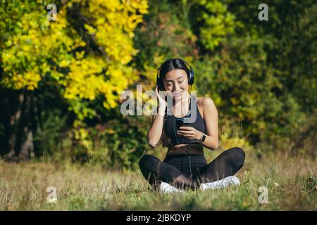 Giovane bella donna asiatica seduta in lotus posizione nel parco, rilassarsi dopo il fitness e la corsa, ascoltare la musica con le cuffie, e utilizzando mob Foto Stock