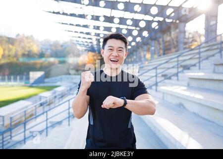 L'atleta maschile felice e di successo guarda la macchina fotografica e sorride, asiatico felice con il risultato del suo allenamento vicino allo stadio al di fuori della mattina Foto Stock