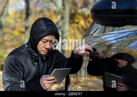 maschio bandito ladro auto ladro asiatico utilizza un tablet per disattivare l'allarme auto Foto Stock