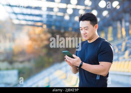 Un uomo sano guarda un video su un telefono cellulare dopo un allenamento mentre si è seduti all'aperto utilizzando un'app su un dispositivo wireless 4g. L'atleta asiatico è felice Foto Stock