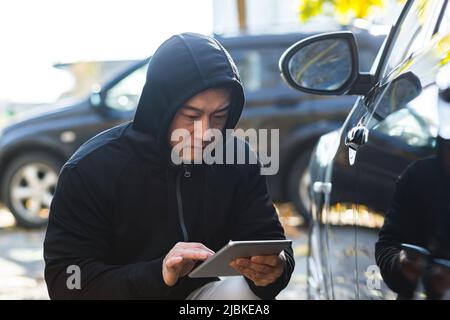 maschio bandito ladro auto ladro asiatico utilizza un tablet per disattivare l'allarme auto Foto Stock