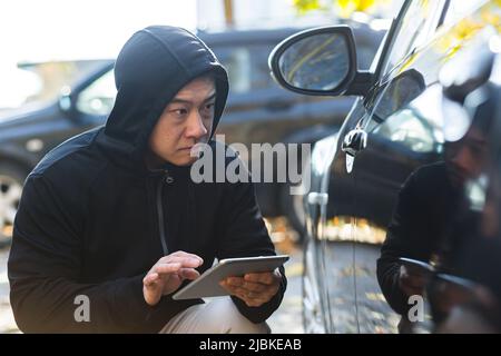 maschio bandito ladro auto ladro asiatico utilizza un tablet per disattivare l'allarme auto Foto Stock