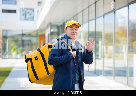 L'uomo di consegna del corriere ambulante asiatico sorridente e guardando la macchina fotografica, ha un grande zaino giallo per la consegna del cibo Foto Stock