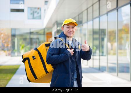 L'uomo asiatico in un cappuccio giallo e con una borsa per la consegna sorrisi guarda la fotocamera e mostra un pollice in su Foto Stock