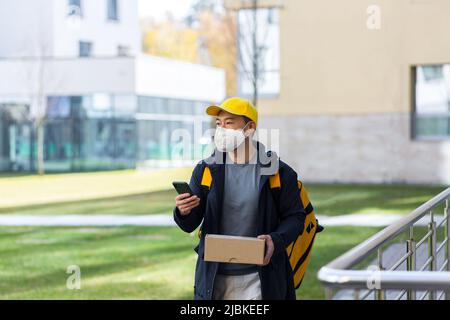 Corriere a piedi in maschera medica di protezione sul fronte di consegna uomo asiatico sorridente alla ricerca di indirizzo di consegna al telefono, ha grande zaino giallo per il cibo Foto Stock