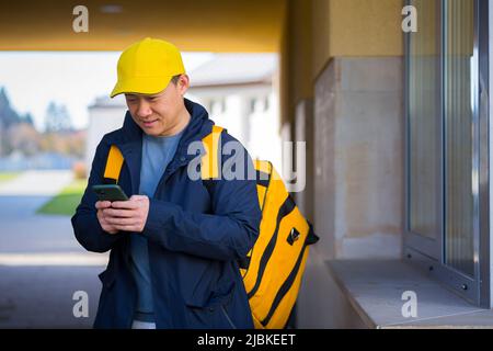 corriere alimentare maschile asiatico utilizzando il cellulare utilizzando l'app per telefono cellulare sullo smartphone, mentre si trova in città strada con isolamento con zaino termico consegna f Foto Stock
