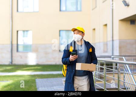 Corriere alimentare maschile asiatico in uniforme gialla utilizzando l'app mobile su smartphone mentre si cammina lungo la strada della città con isolamento con zaino termico deliverin Foto Stock