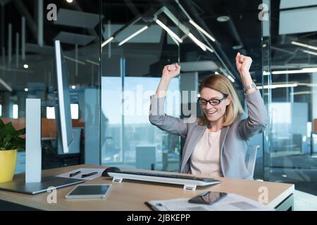 Bella e di successo donna d'affari con occhiali, celebrando la vittoria guarda lo schermo del monitor del computer e si rallegra nel comple con successo Foto Stock