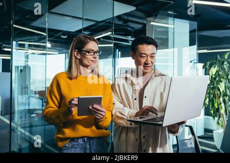 Incontro d'affari di due dipendenti, colleghi asiatici e femminili, discutere il progetto, lavorare in un ufficio moderno, guardare uno schermo di laptop, discus Foto Stock