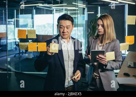 Due dipendenti, scrivendo un piano e compiti sul task board, un uomo asiatico e una donna, lavorando in un ufficio moderno, scrivono compiti su note colorate Foto Stock