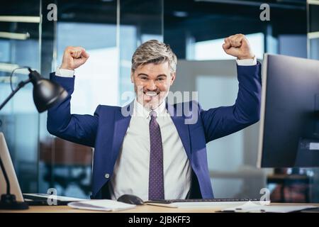 Il boss di successo lavora in un ufficio moderno al computer, un uomo anziano grigio e felice con una barba guarda la fotocamera e sorride, tiene le mani in su, Foto Stock