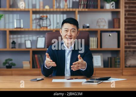 Uomo d'affari allegro guardando la webcam, sorridendo e gioendo gesto volentieri mani, conferenza online riunione, successo asiatico Foto Stock